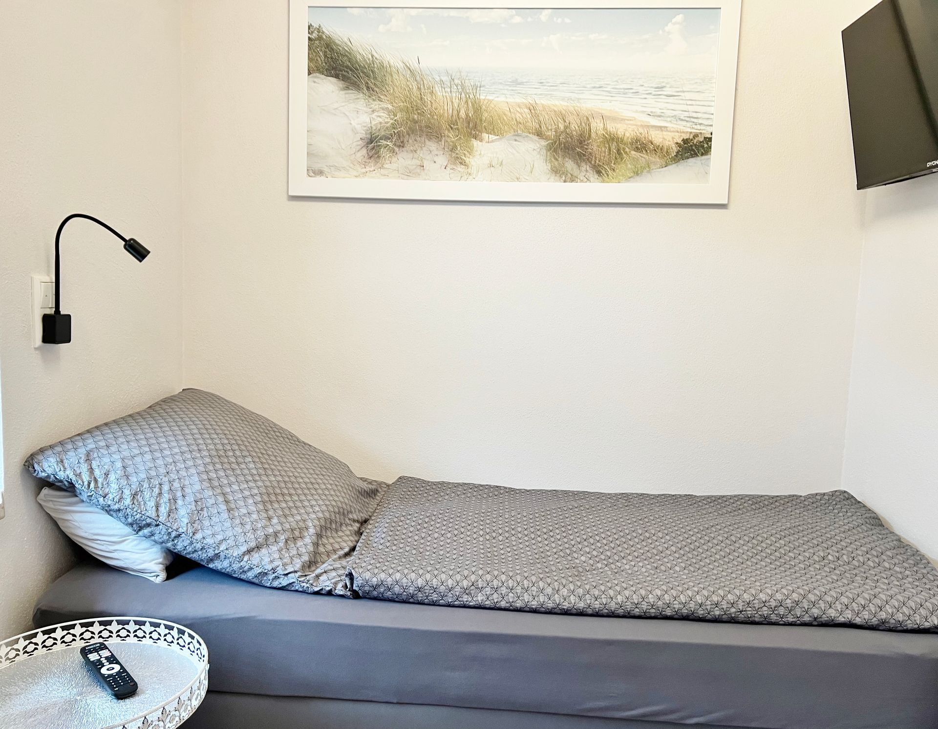 Ein Einzelbett mit grauer Bettwäsche, eine an der Wand befestigte Leselampe und ein Gemälde einer Küstenlandschaft in einem kleinen, weißen Zimmer.