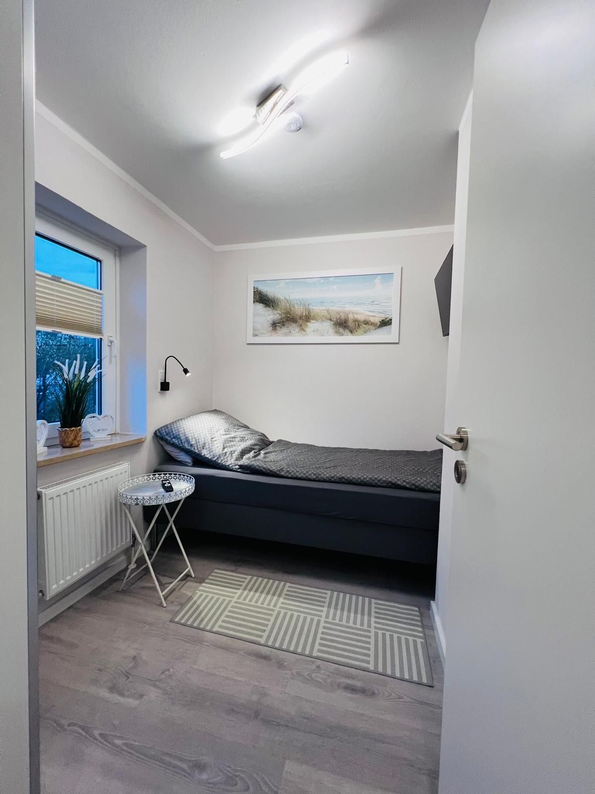 Ein kleines, minimalistisches Schlafzimmer mit einem Einzelbett mit grauer Bettwäsche, einem Beistelltisch, einem Fenster und Wandbildern.