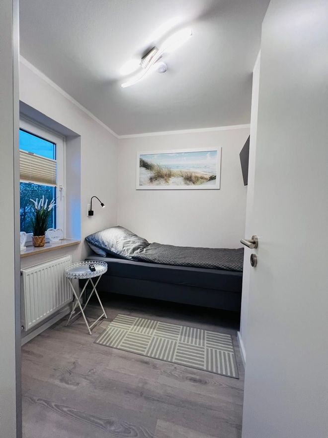 Ein kleines, minimalistisches Schlafzimmer mit einem Einzelbett mit grauer Bettwäsche, einem Beistelltisch, einem Fenster und Wandbildern.