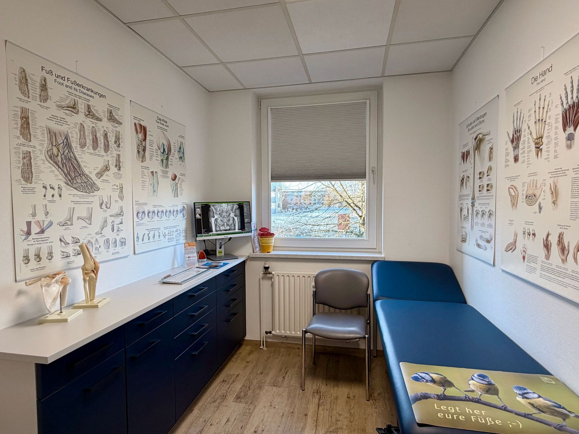 Untersuchungsraum mit Krankenakten, blauen Schränken, Untersuchungstisch, Fenster.