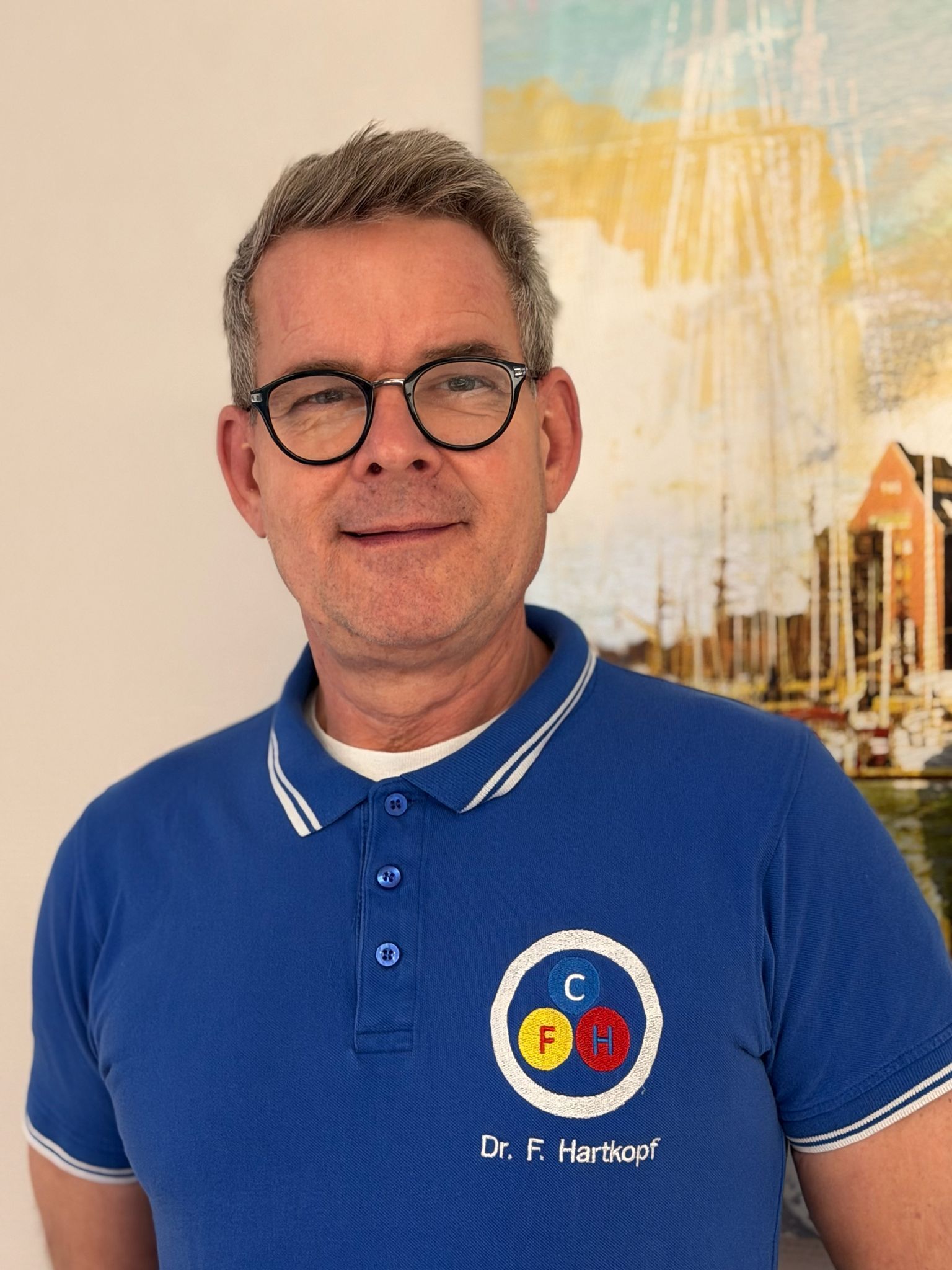 Ein Mann mit Brille und blauem Poloshirt mit Logo lächelt und steht vor einem Gemälde.