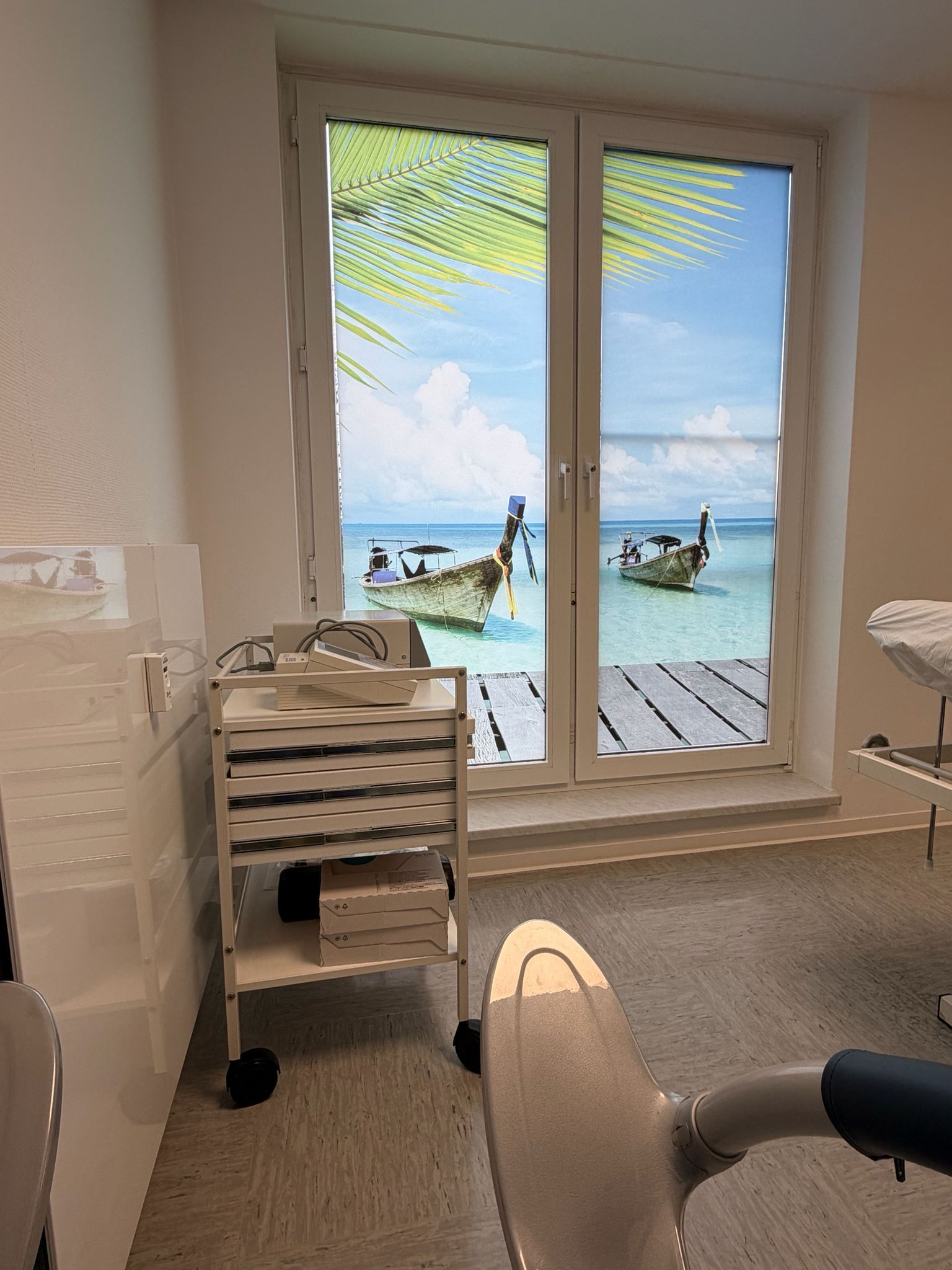 Innenansicht eines Zimmers mit einem Fenster, das einen Blick auf eine Strandszene bietet. Ein Rollwagen und ein Stuhl befinden sich im Zimmer.