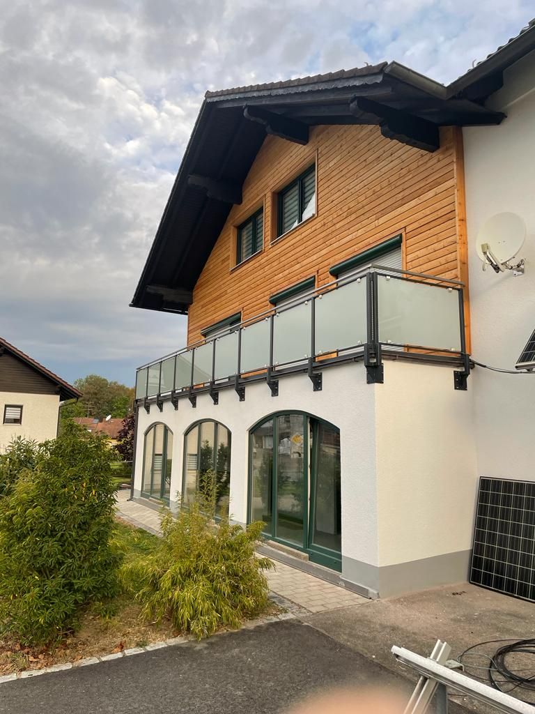 Zweistöckiges Haus mit hölzernem Obergeschoss, Balkon und Bogenfenstern. Grüne Tür, bewölkter Himmel.
