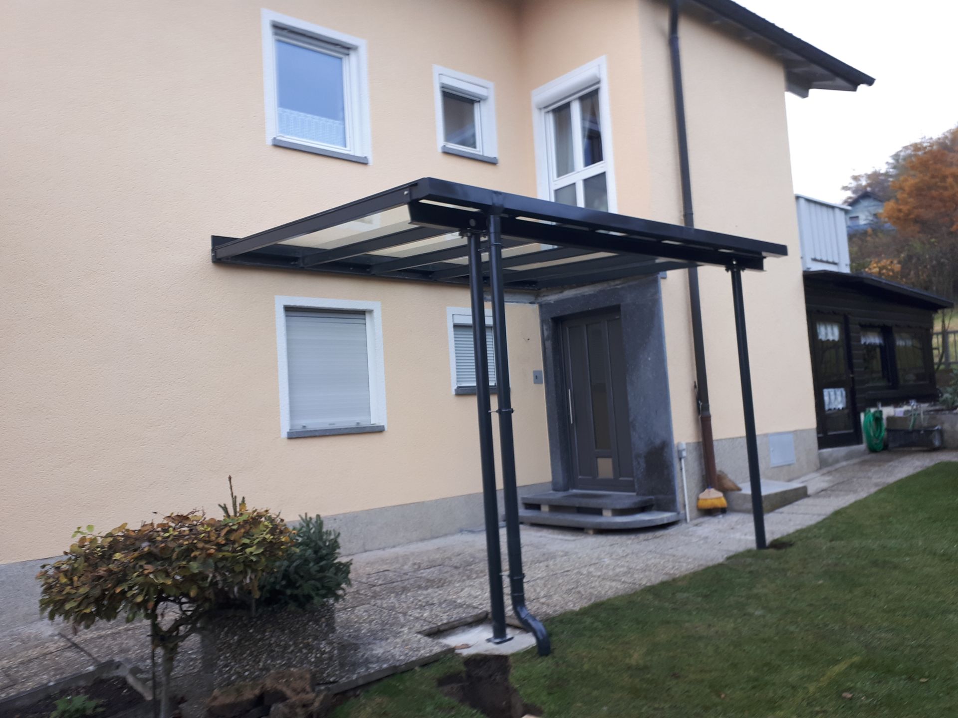 Ein überdachter Eingang zu einem beigefarbenen Haus mit schwarzem Vordach und Stützbalken.