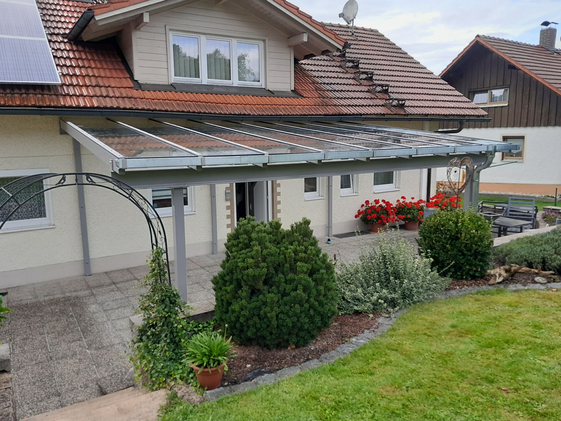 Haus mit überdachter Terrasse, Garten und rotem Ziegeldach. Im Vordergrund grüner Rasen und Sträucher.