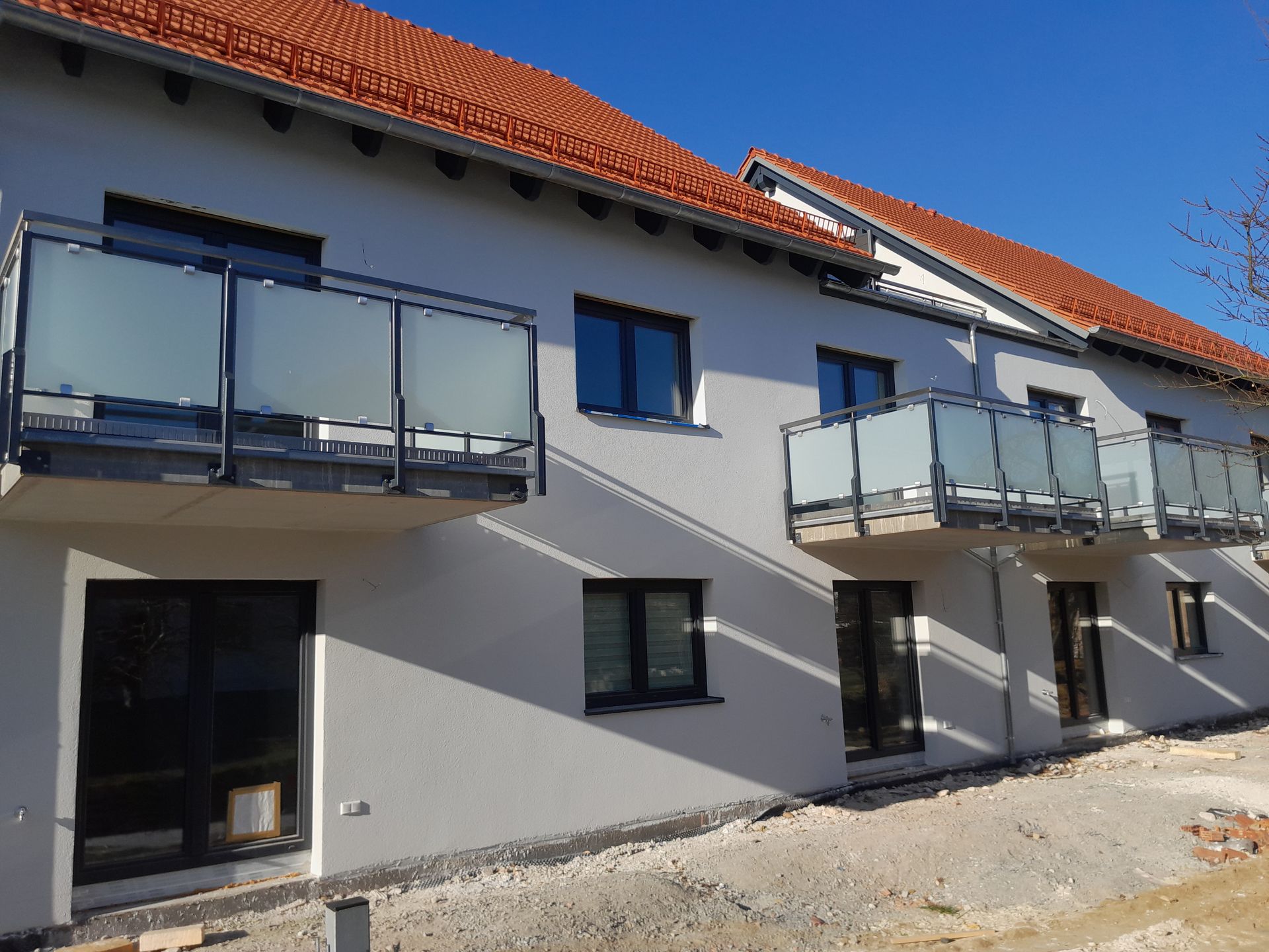 Zweistöckiges Gebäude mit rotem Ziegeldach, Balkonen mit Milchglas und schwarzen Fensterrahmen unter klarem, blauem Himmel.