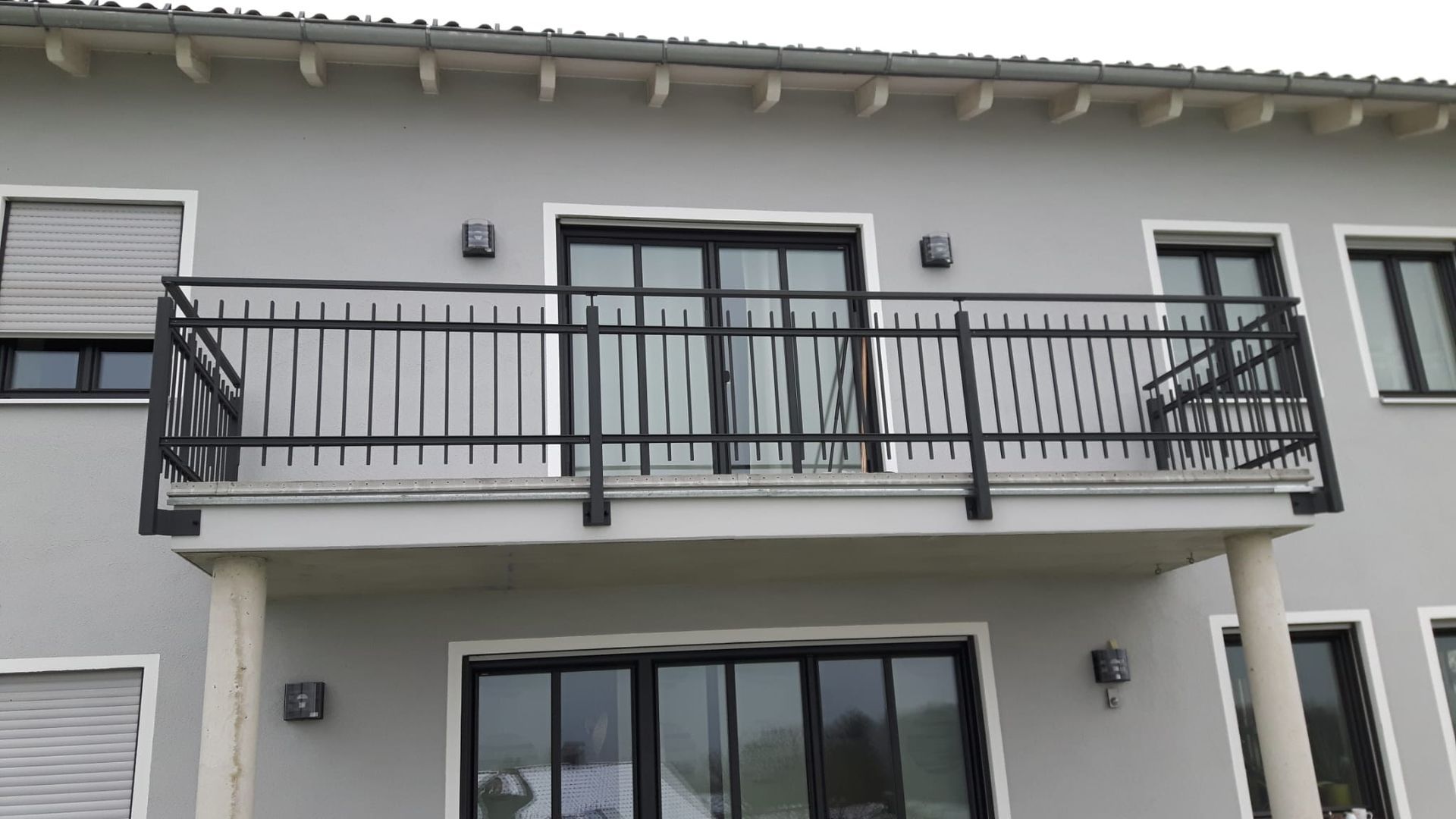 Außenansicht eines grauen Hauses mit Balkon, der über schwarze Metallgeländer und große Glasschiebetüren verfügt.