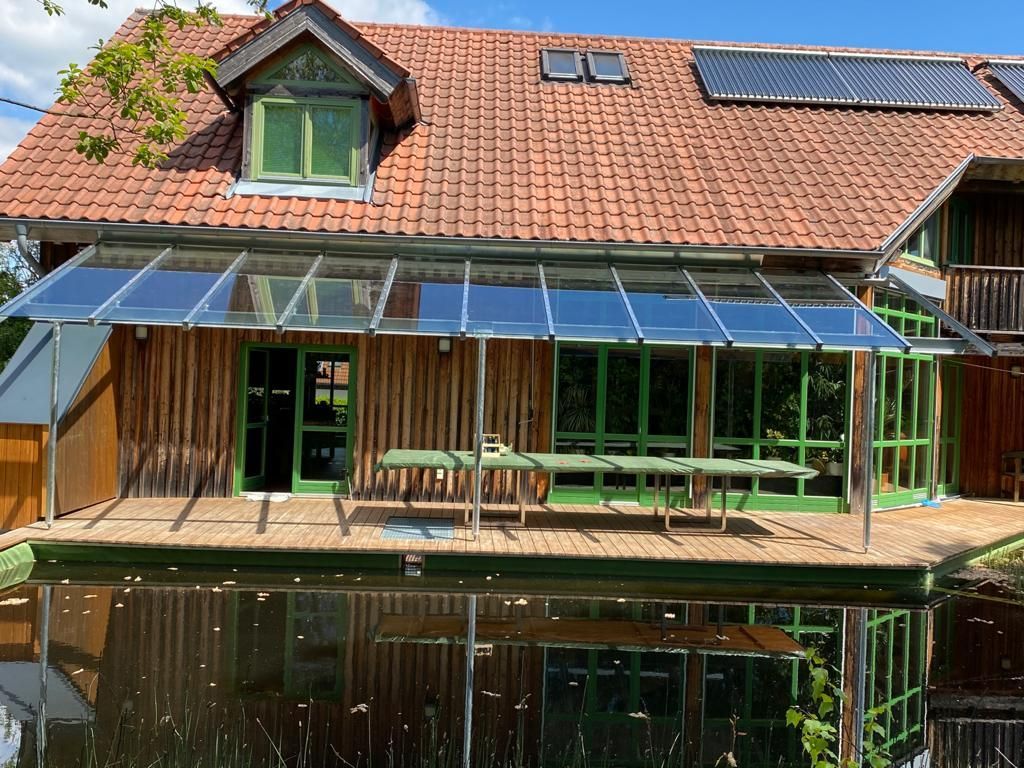 Holzhaus mit verglaster Veranda, die sich im ruhigen Wasser spiegelt. Grüne Türen und Fenster; rotes Ziegeldach.