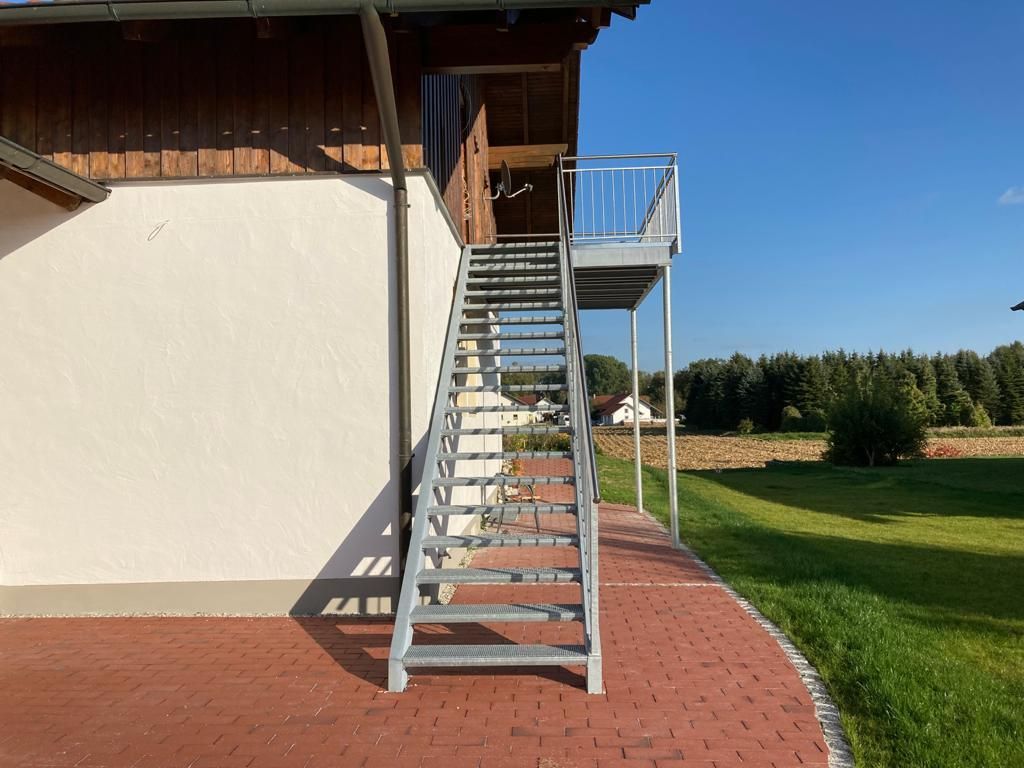 Eine metallene Außentreppe führt zu einem Balkon, der an ein Gebäude angebaut ist und sich vor blauem Himmel abhebt.