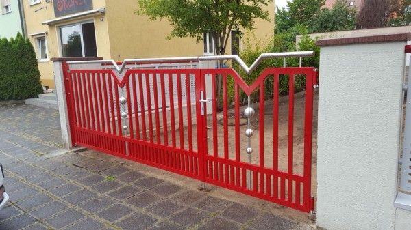Ein modernes, zweiflügeliges Einfahrtstor mit roten, vertikalen Metallstäben, verziert mit dekorativen silbernen Kugeln und oberen Akzenten.