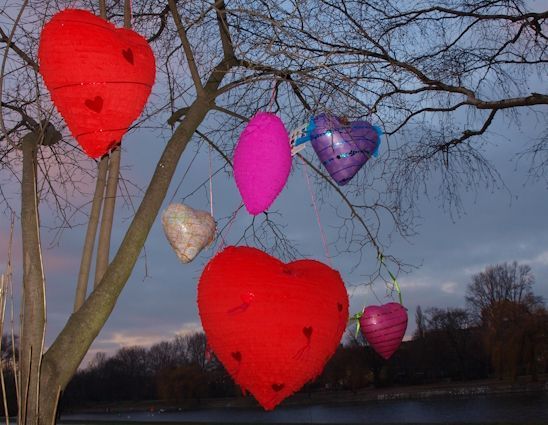 Herzen in verschiedenen Farben und Formen hängen an einem Ast vor einem dämmerigen Himmel.