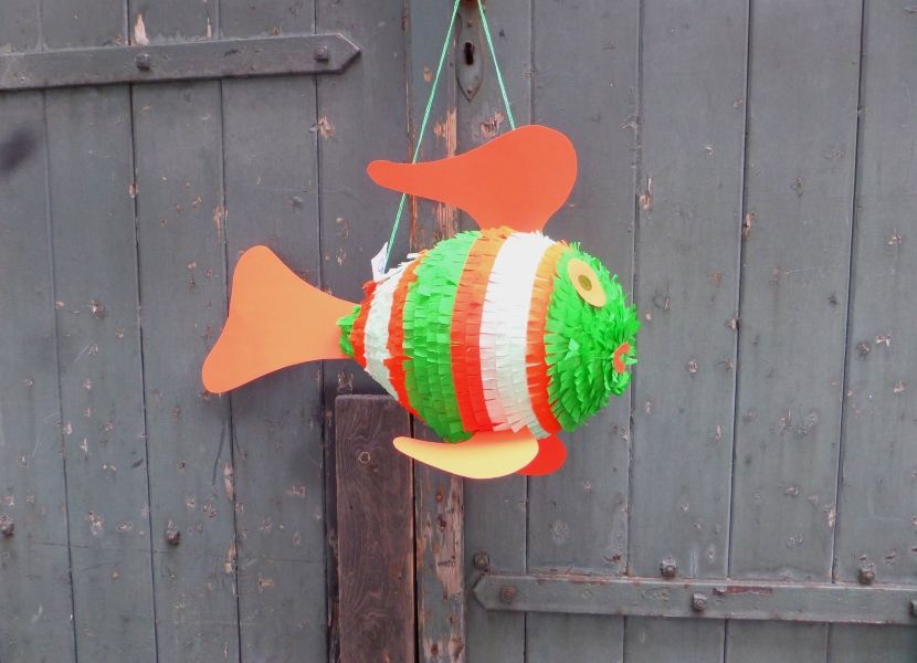 Eine fischförmige Piñata in Grün, Weiß und Orange hängt an einer verwitterten Holztür.
