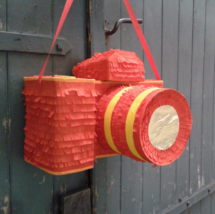 Eine Kamera-Piñata aus rotem und gelbem Fransenpapier hängt an einer Tür.