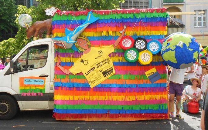 Festwagen mit Regenbogenfransen, olympischen Ringen, Globus und einer Person, die ein Schild hält.