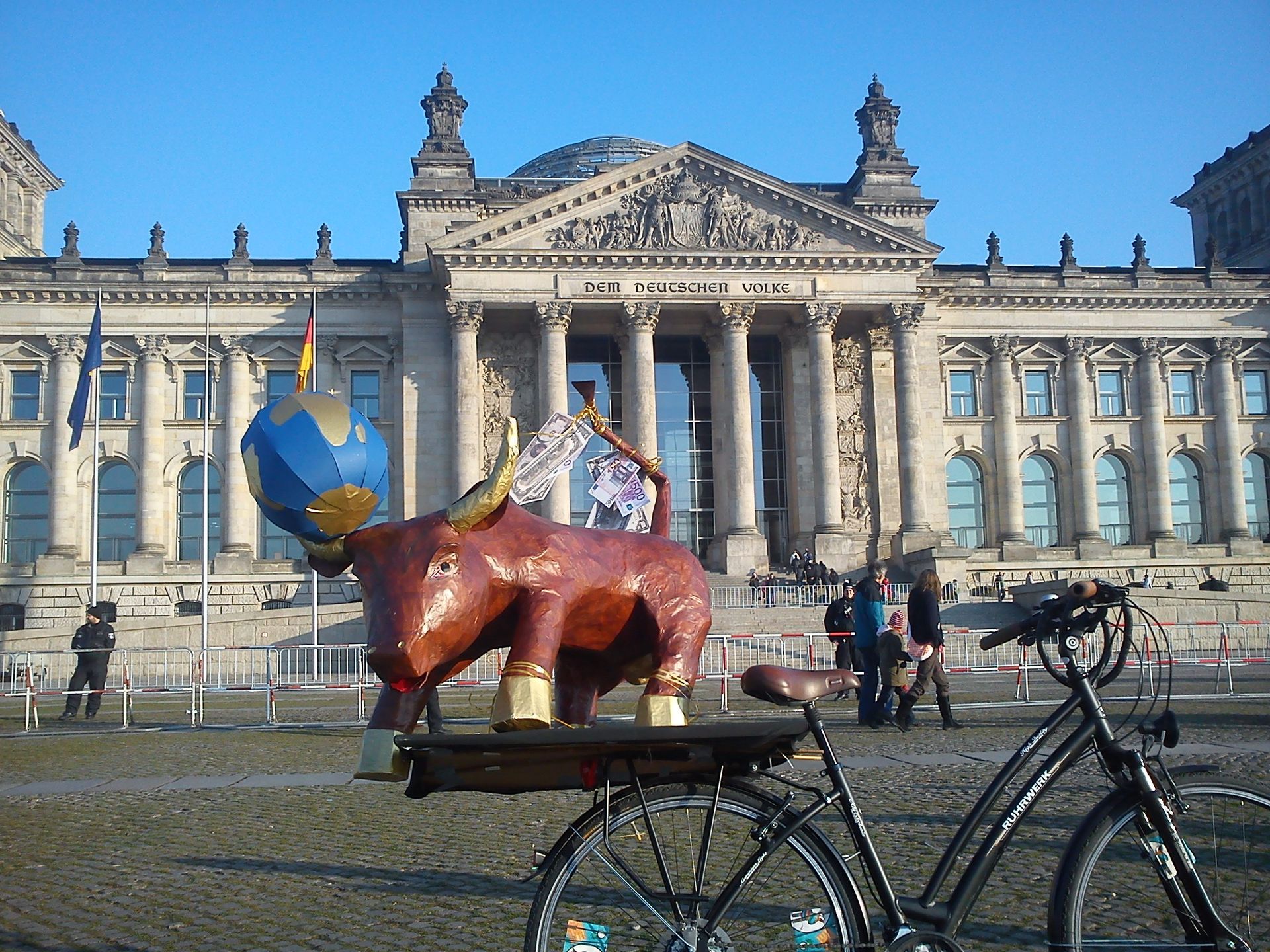 Ein Fahrrad vor dem Reichstagsgebäude in Berlin, neben dem eine rote, stierähnliche Skulptur einen Globus hält.
