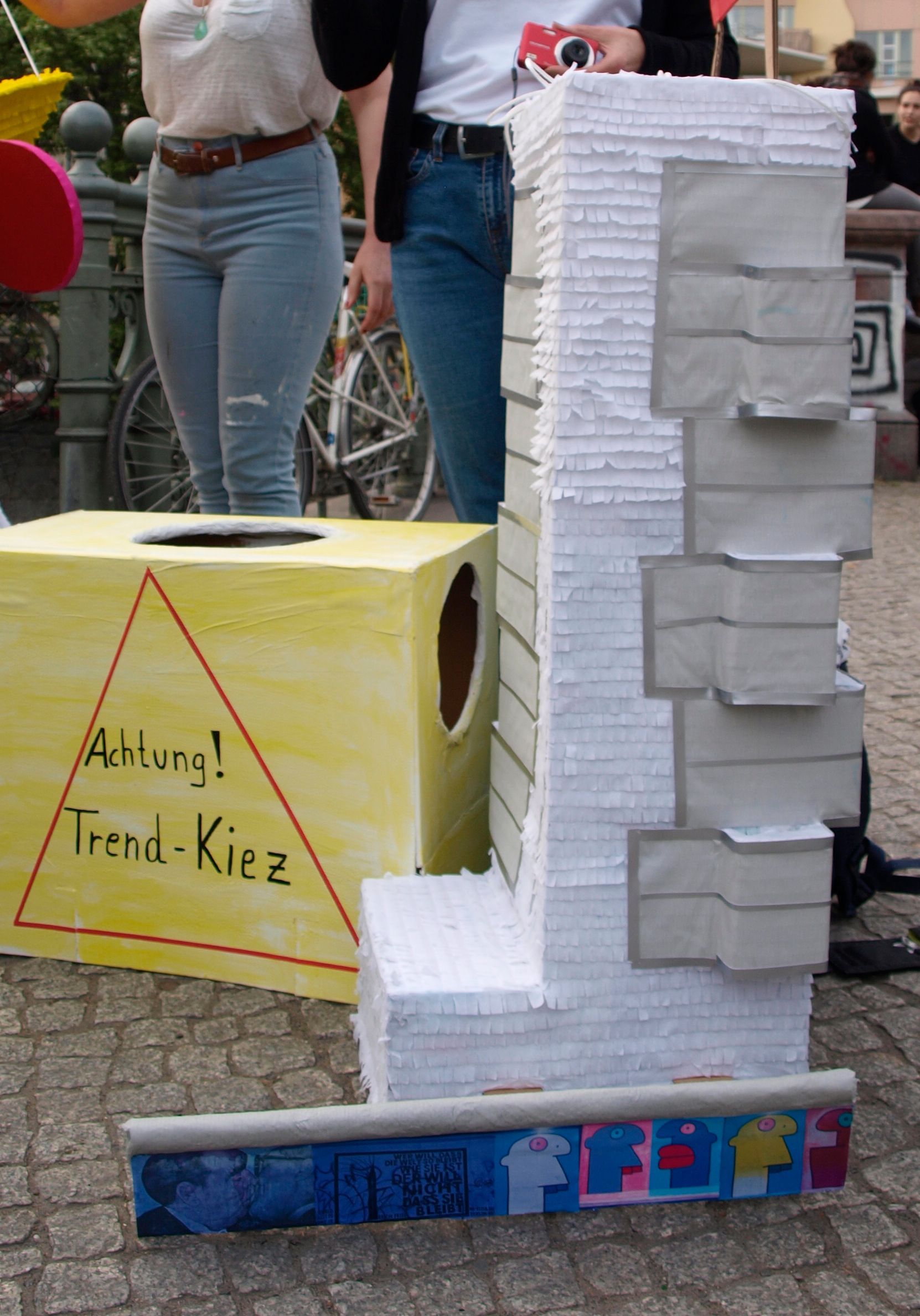 Eine hohe, L-förmige Skulptur aus Papier steht neben einer gelben Kiste. Menschen stehen in der Nähe.