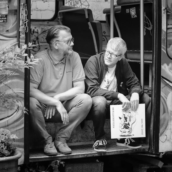 Zwei Männer sitzen auf den Stufen eines Nahverkehrsbusses und schauen nach rechts und links. Einer der beiden Männer hält lässig eine Schallplatte der Band MetzgerButcher in der Hand.