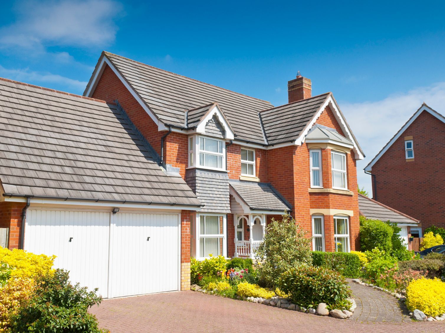 Backsteinhaus mit grauem Dach, weißen Garagentoren und farbenfroher Landschaftsgestaltung unter einem klaren blauen Himmel.