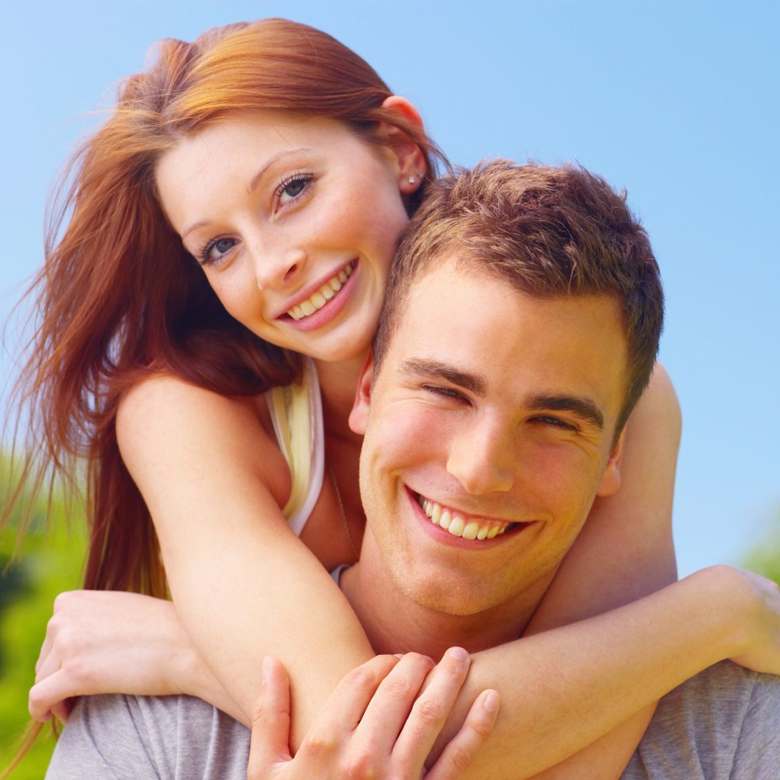 Lächelndes junges Paar, Frau mit roten Haaren umarmt Mann im Freien, blauer Himmel.
