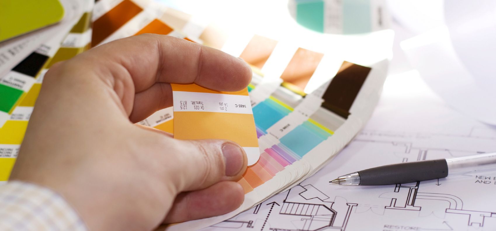 Hand hält ein Farbmuster mit Farbpalettenfächer, Architekturplänen und einem Stift.