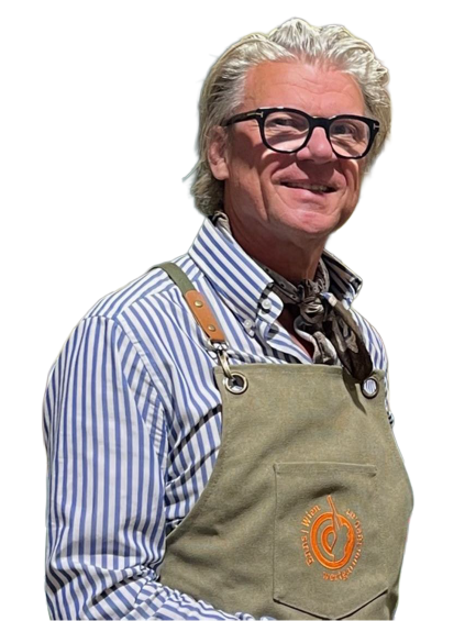 Eine lächelnde Person mit welligem Haar und Brille, die ein gestreiftes Hemd, einen Halsschal und eine hellbraune Markenschürze trägt.
