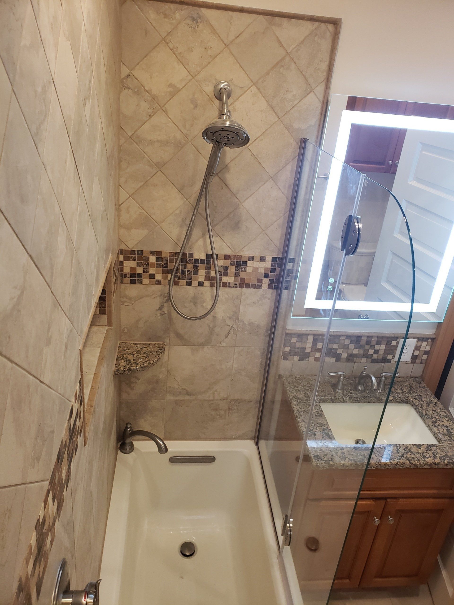 View of bathtub, tiled walls, stationary glass panel for shower door, corner shower shelf, sink, granite sink countertop, mosaic tile backsplash, vanity and mirror with lighting in Cheltenham bathroom remodel. 