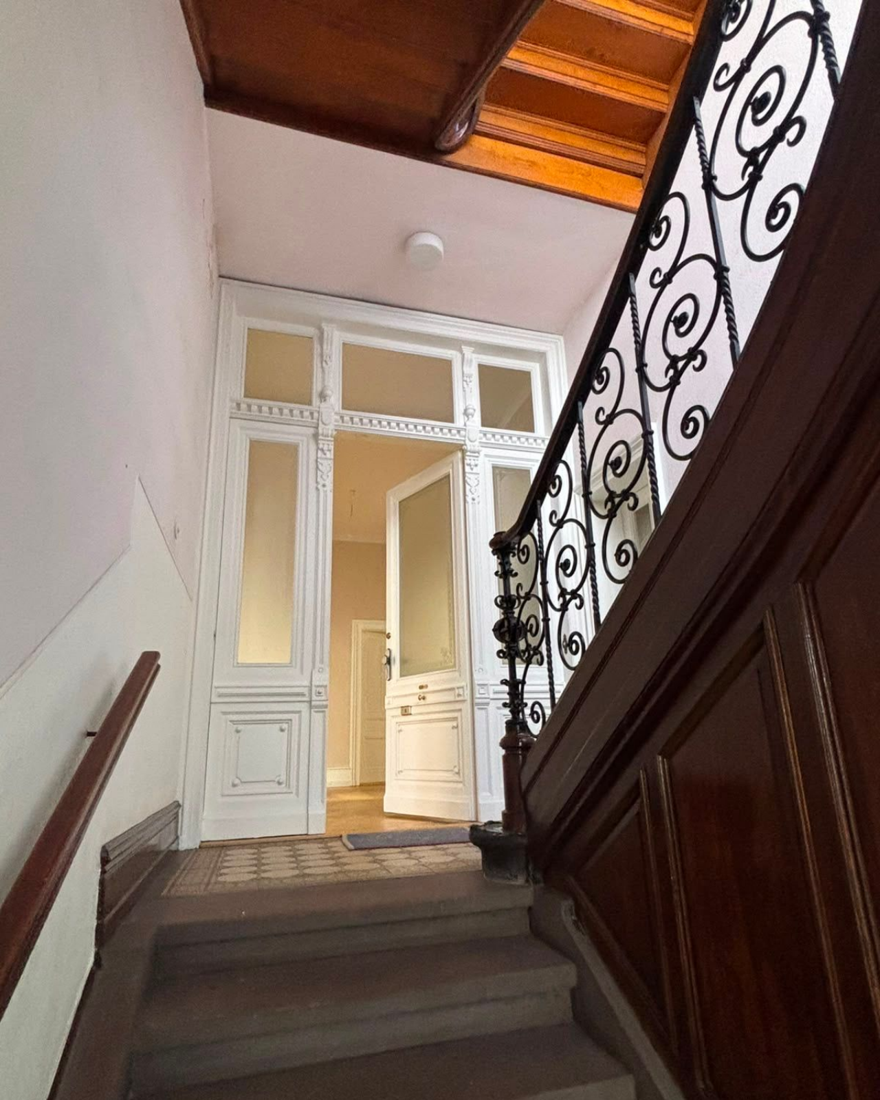 Altbau Treppenhaus. Weiße Eingangstür. Braune Holztreppe. Metallgeländer mit Schnörkeln.
