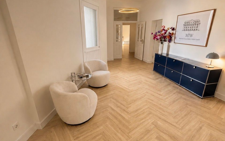 Eingangsbereich. Fischgrätparkett. Zwei beige Sessel. kleiner runder Tisch. Blaues USM Haller Möbel. Bild mit MIW Logo.