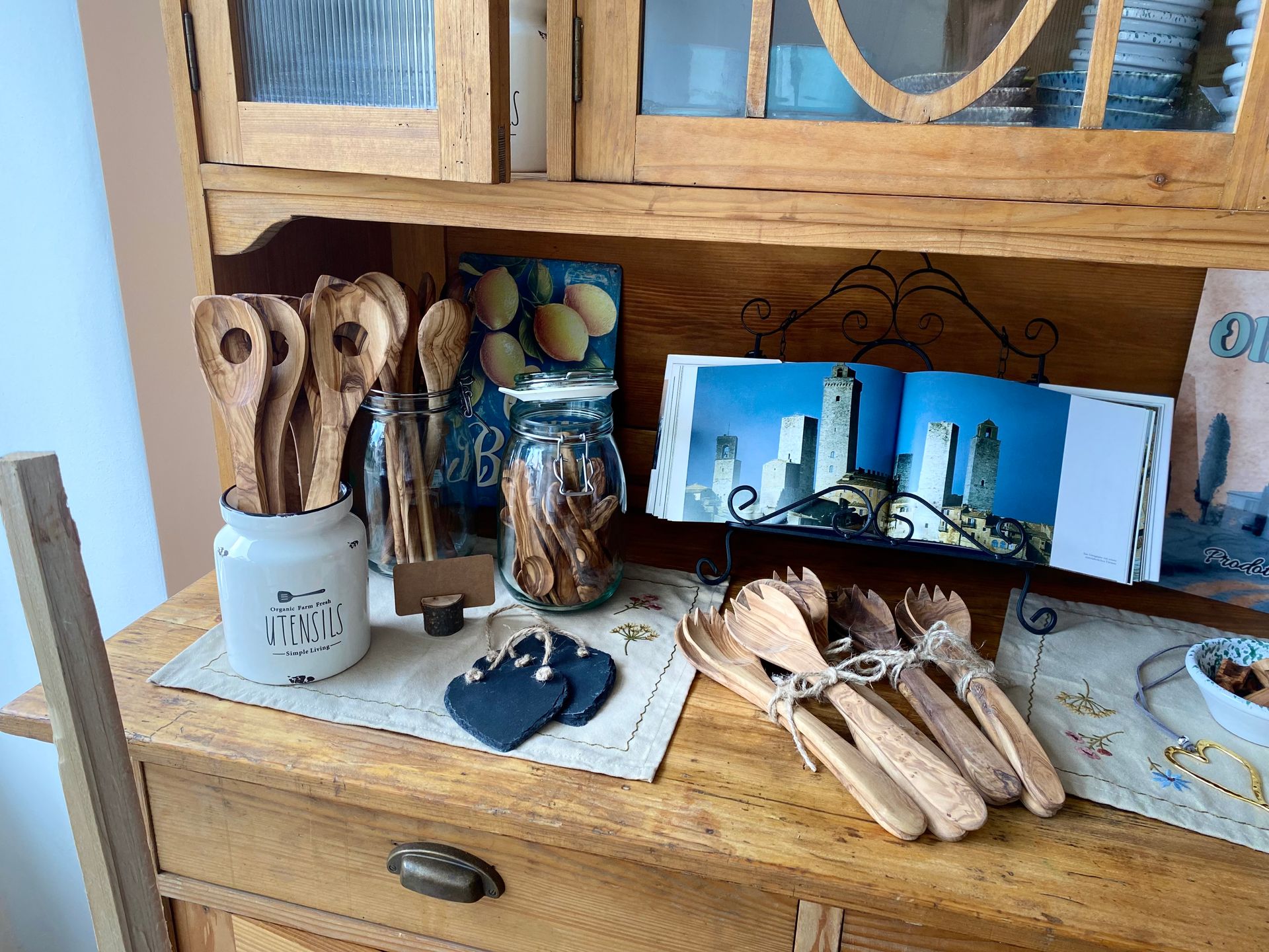 Holzregal auf dem ein offenes Buch mit italienischen Bildern neben einigen Olivenholzprodukten drapiert ist 