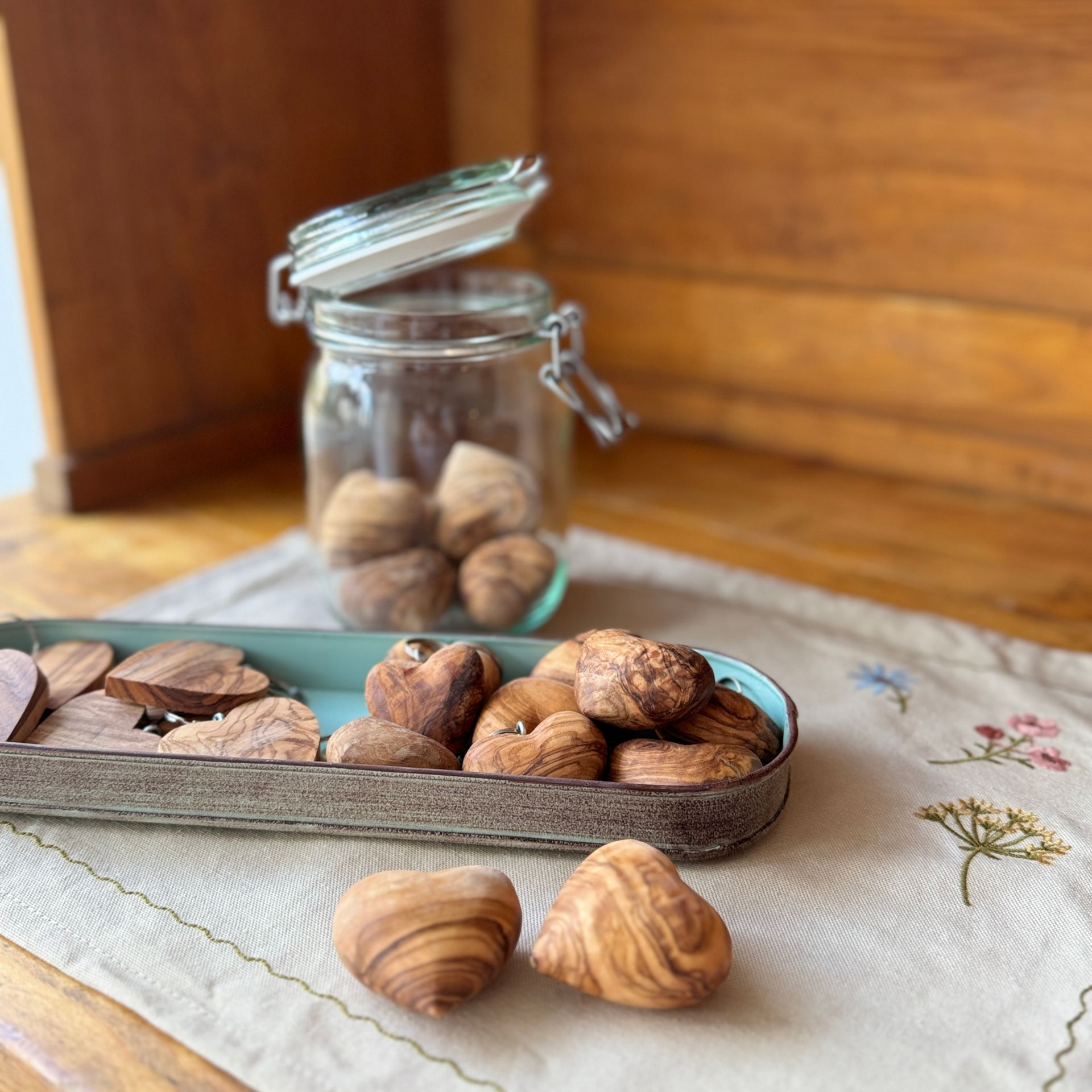 Mehrere herzförmige Olivenholzstücke auf Leinentuch, daneben ein Glasgefäß auf einem Holztisch