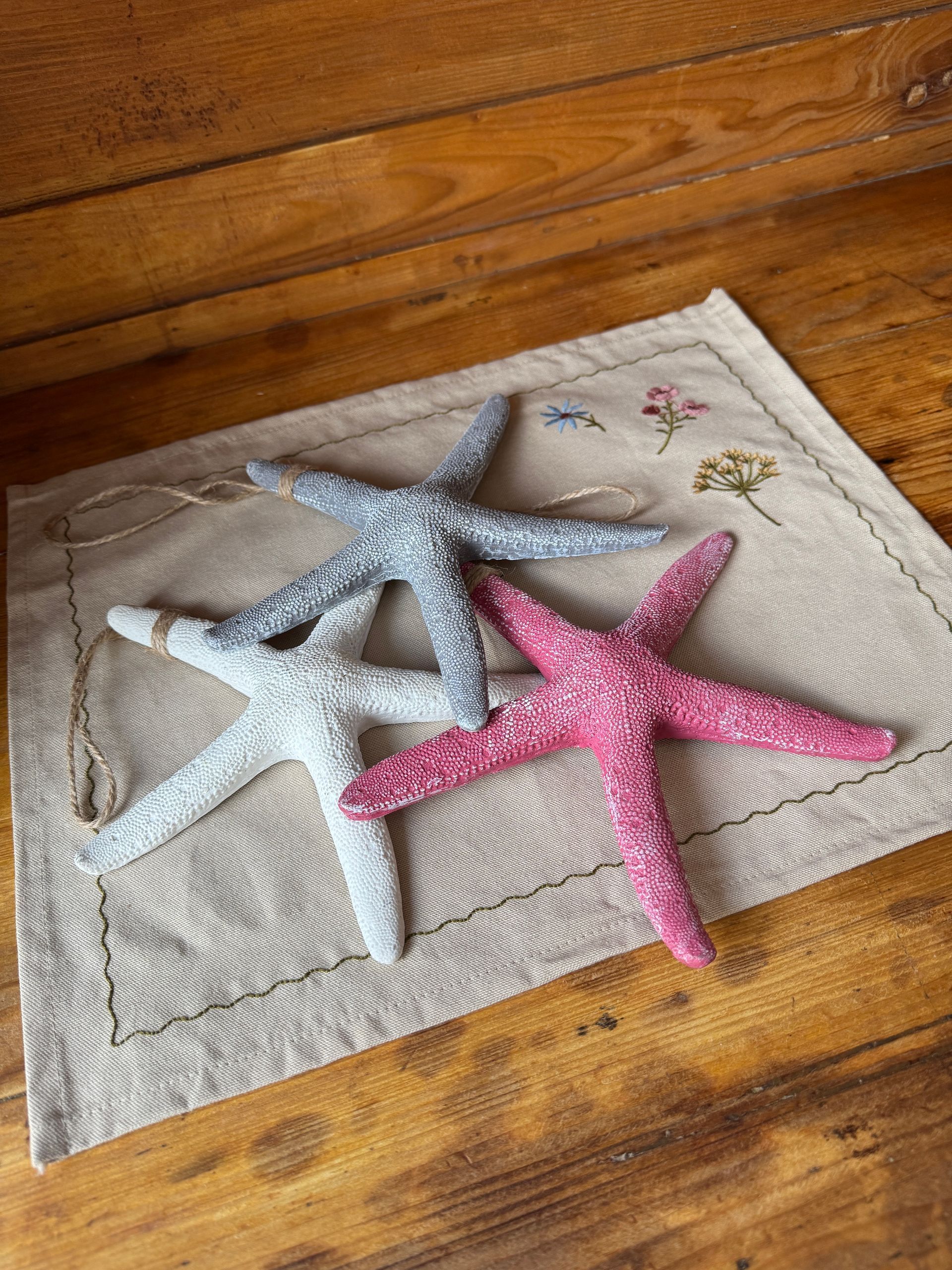 Seesternanhänger in grau, pink und weiß auf einer mit kleinen Blumen bestickten, naturfarbenen Stoffserviette