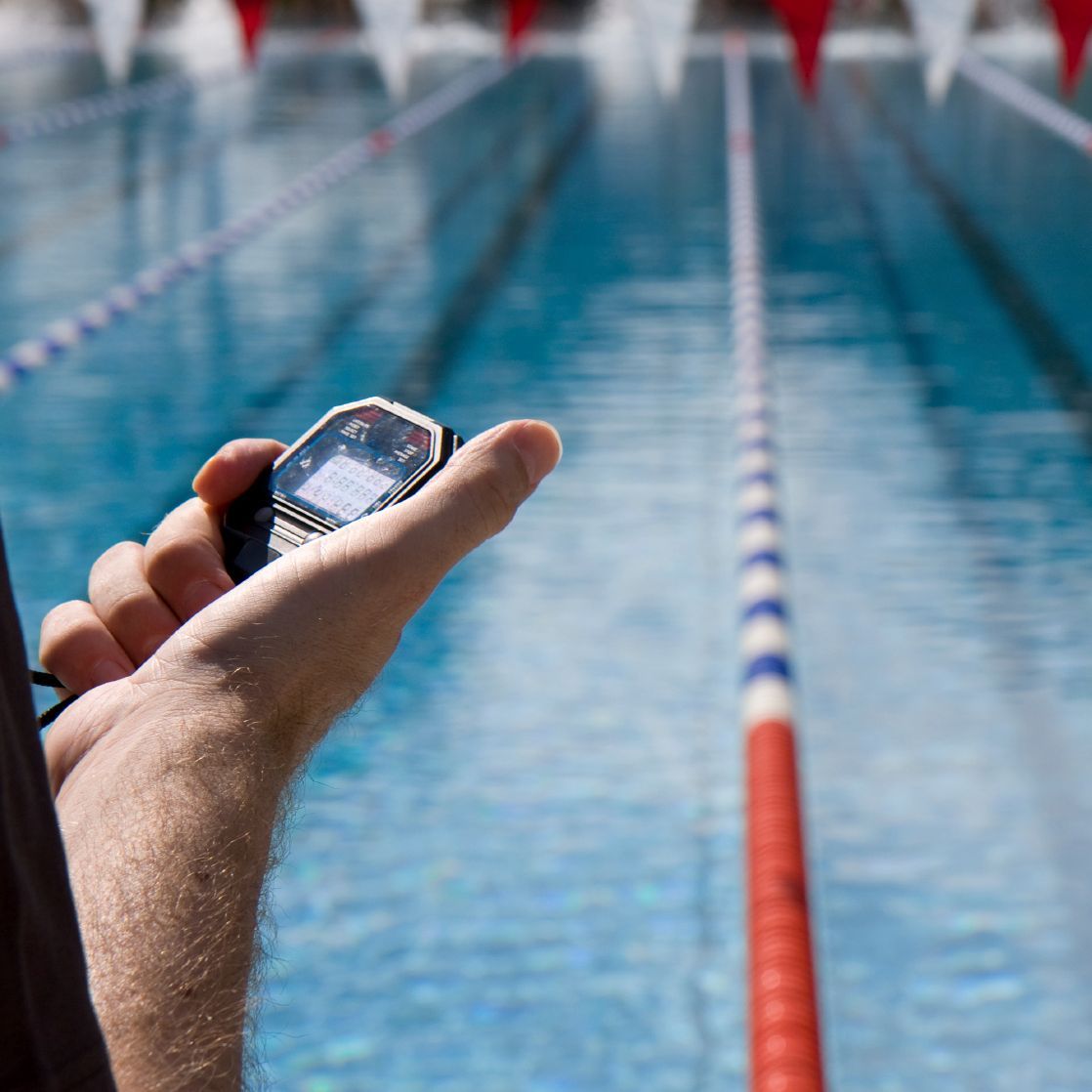 Eine Hand hält eine Stoppuhr vor dem Schwimmbecken