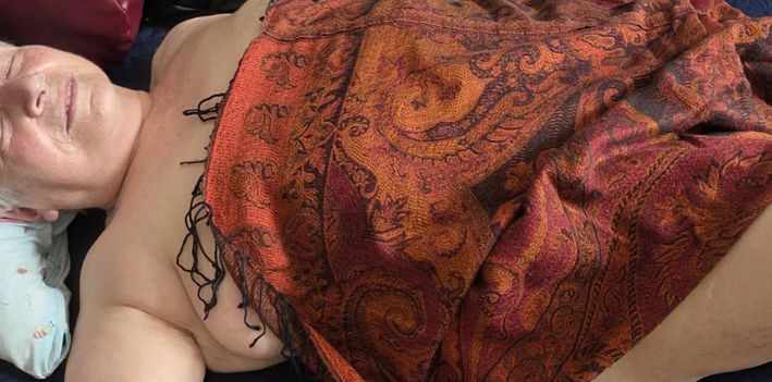 eine ältere Frau liegt mit einem Tuch in warmen Farben bedeckt . Sie hat die Augen geschlossen und lächelt seelig - Symbol für daie Wirkung von Sexualassistenz bei Demenz.
