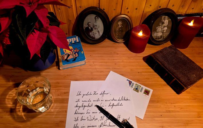 Auf einem Holztisch stehen an der Wand eine Topfblume,zwei Kerzen, gerahmte alte Fotografien. Auf dem Tisch selber liegen zwei vom Gebrauch abgenutzte Bücher - das eine ist 