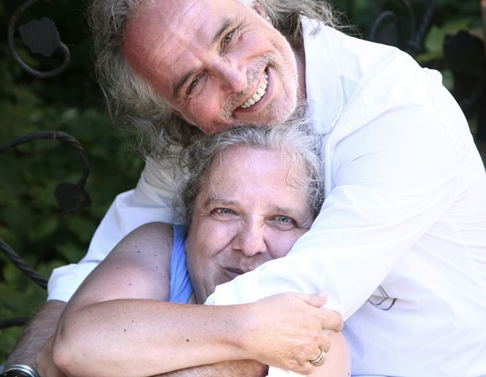 Eine ältere Frau hat sich in den Arm eines Mannes im weißen Hemd gekuschelt und schaut verschmitzt lächelnd in die Kamera - Symbol für  Nähe und Gebborgenheit in den Armen des Sexualbegleiters