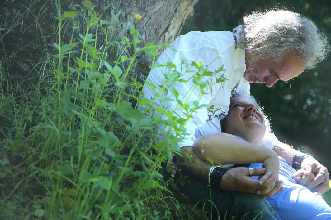 im Schatten eines Baumes sitzt im grünen Gras ein älterer Mann, Sexualbegleiter, und auf seinem Schoß liegt eine behinderte Frau. Sie halten sich bei den Händen und die Frau strahlt ihn an - als Sinnbild für Symbol für Wachstum, Würde und reflektierte Moral