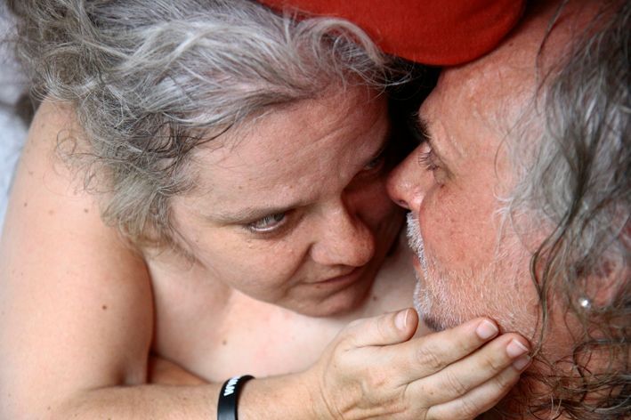 eine ältere, grauhaarige Frau ohne sichtbare Kleidung kuschelt sich an einen grauhaarigen Mann, blickt ihn an und streichelt sein Gesicht - Symbol für Intimität bei der Sexualbegleitung von dementen Frauen