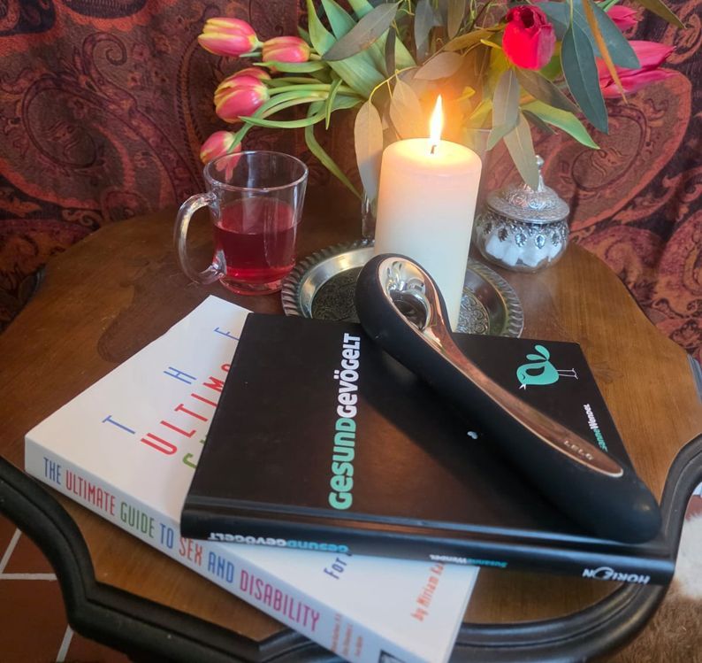 auf einem Tiscchen liegen vor einer Blumenvase mit Tulpen, einer Tasse Tee und einer brennenden Kerze zwei Bücher: