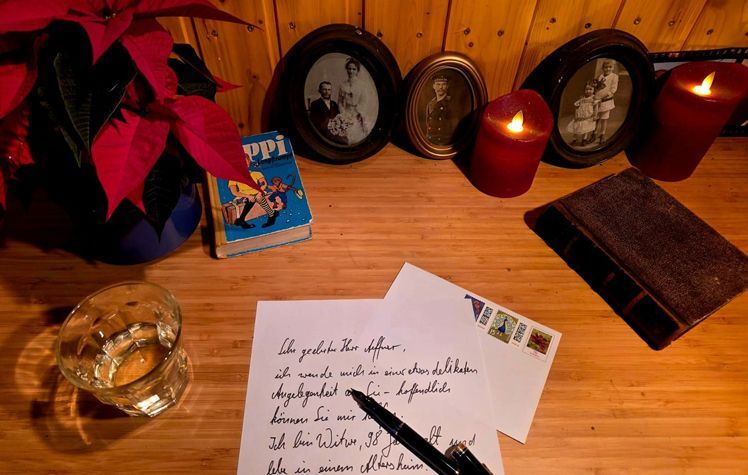 Auf einem Schreibtisch stehen eine Pflanze, 2 Kerzen, alte gerahmte Fotos, ein Glas Wasser, ein altes Buch, ein zerlesenes Buch 