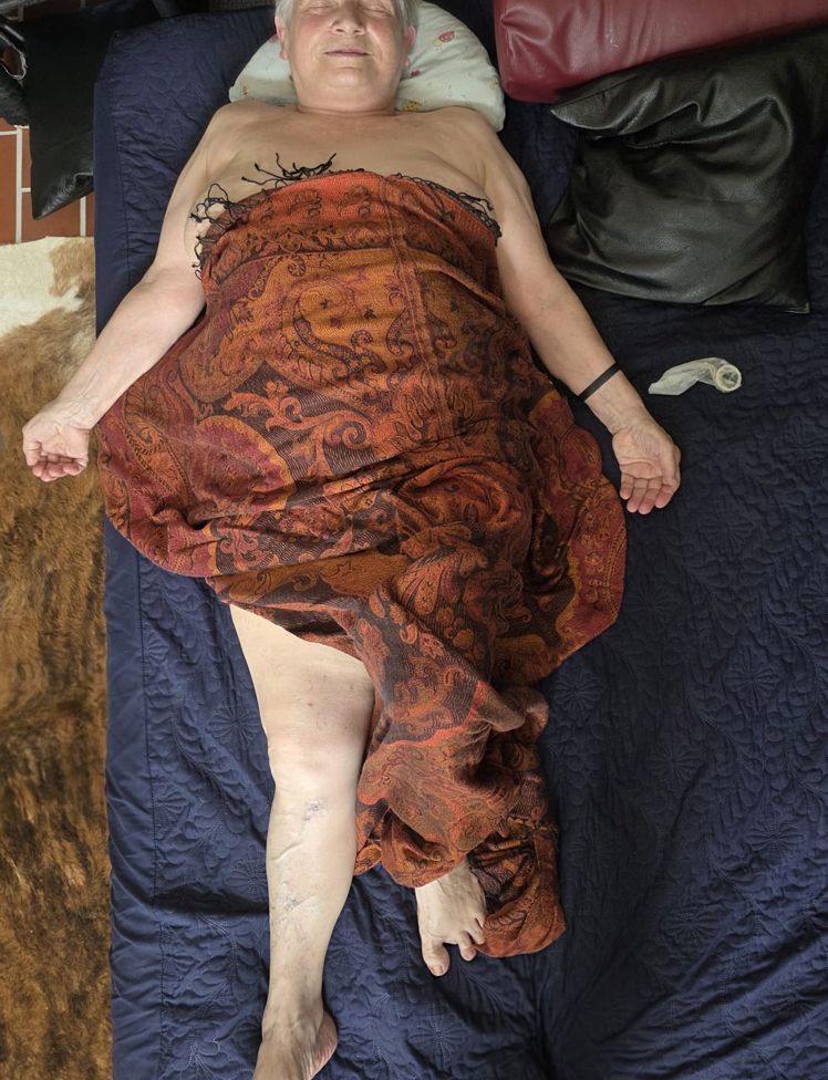 auf einem Bett mit Kissen liegt mit entspanntem, zufriedenem Gesichtsausdruck eine alte Frau, mit einem schönen Tuch mit warmen Farben bedeckt, daneben ein gebrauchtes Kondom - als Symbol für mehr Lebensqualität durch Sexualbegleitung im Alter