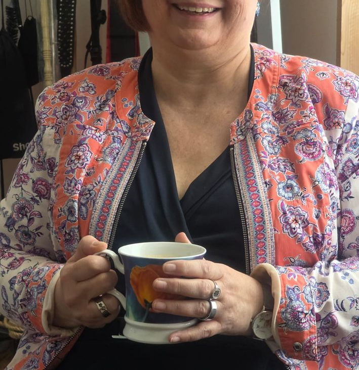 eine ältere Dame mit einer bunten, farbenfrohen Jacke hält eine Tasse in den Händen und lächelt - Symbol für mehr Lebensfreude und entspannten Alltag durch Sexualbegleitung