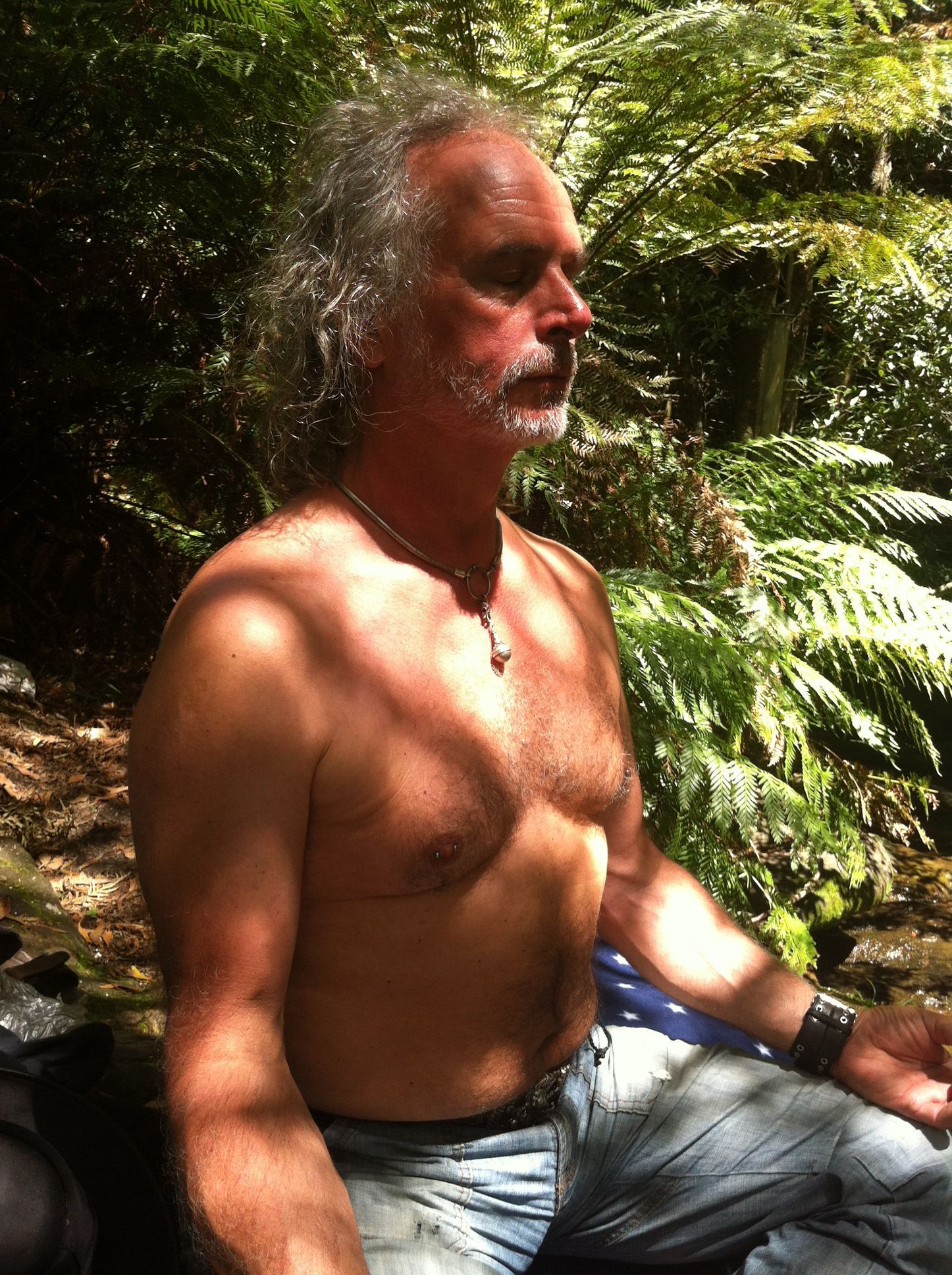 Vor grünem Farn sitzt Thomas Aeffner in Meditationshaltung. Sonnenlicht und Schatten von Bäumen spielen auf der Haut seines nackten Oberkörpers. Symbol dafür, in sich zu ruhen, sein IKIGAI gefunden zu haben.