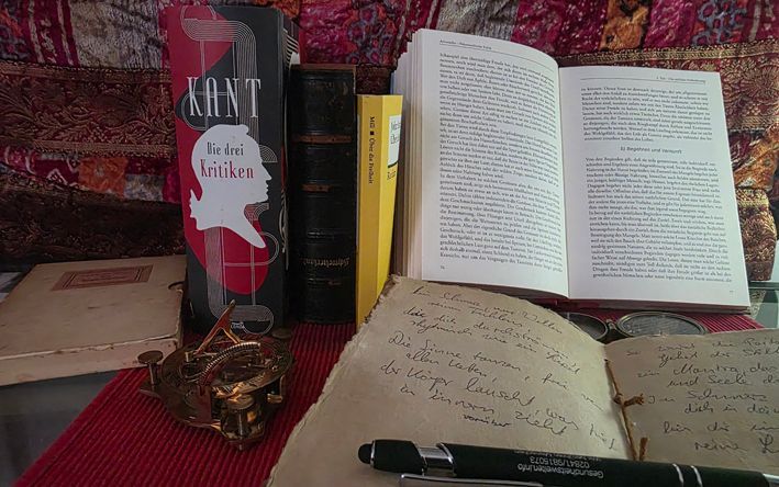 auf einem Schreibtisch stehen und liegen moralphilosophische Bücher : Kant, Mill Arestoteles etc. ein aufgeschlagenes Notizbuch mit einem Stift und ein Nautischer Kompass/Sonnenuhr liegen davor - als Sinnbild dafür, dass wir moralische Postulate hinterfragen sollen, um zu heute passenden Moralvorstellungen zu kommen.