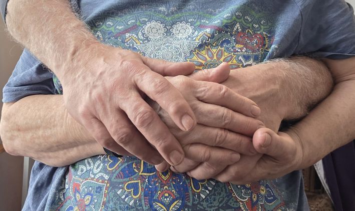 die Hände von einer älteren Frau und einem Mann halten und verschränken sich ineinander vor einer Bluse mit fröhlichem Muster - Symbol für den liebevollen, respektvollen Kontakt von Klientin und Sexualbegleiter