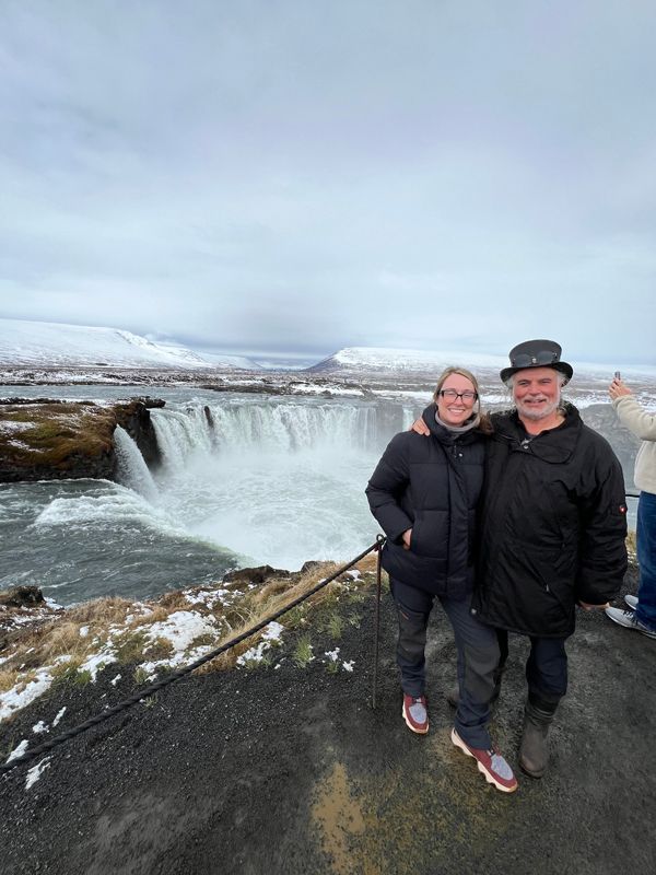 Thomas und seine erwachsene Tochter stehen in Island am Geländer und schauen in die Kamera. Hinter ihnen ist ein Wasserfall zu sehen