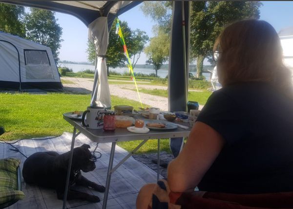 Blick aus dem Vorzelt über ein Stückchen Campingplatz auf den Pilsensee. Im Vordergrund Frau am Frühstückstisch, Hund liegt unter dem Tisch