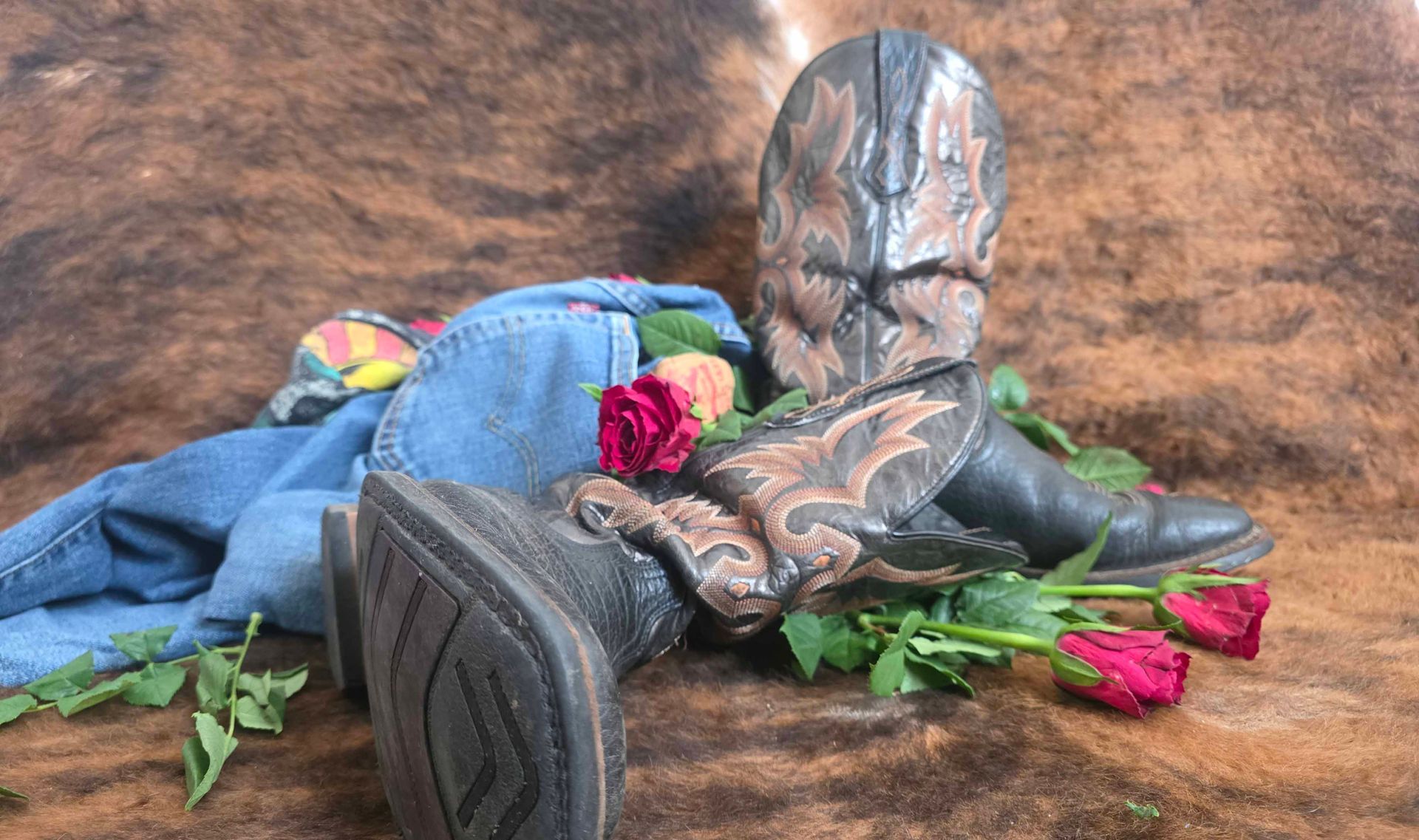 zwei Cowboystiefel, Jeans, T-Shirt und drei rote rosen liegen auf dem Boden