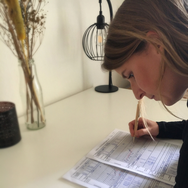Frau über eine Zeichnung gebeugt, beleuchtet von einer Schreibtischlampe. Weißer Schreibtisch, dekorative Vase im Hintergrund.
