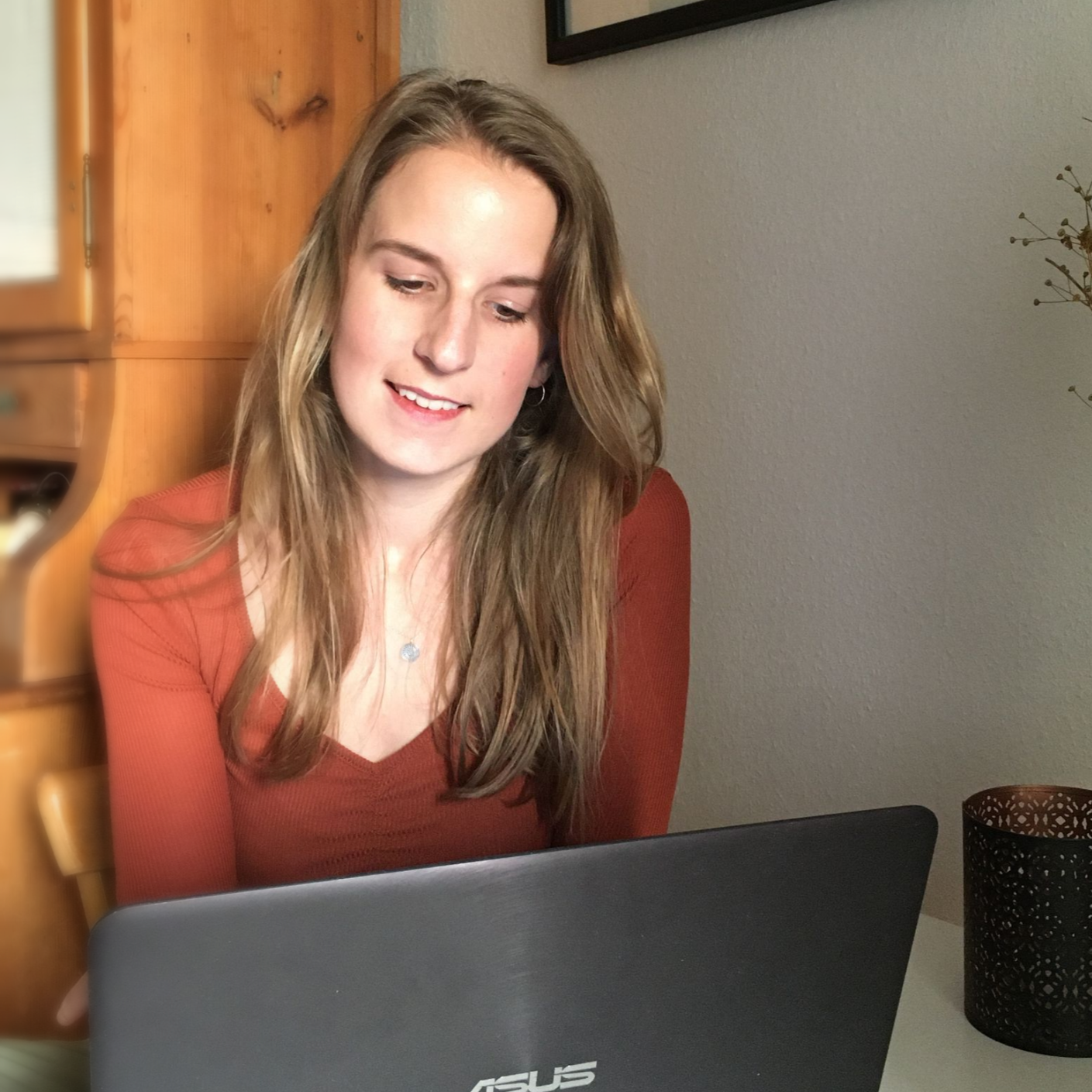 Eine lächelnde Frau arbeitet an einem Tisch an einem Laptop. Sie trägt ein rostfarbenes Hemd. Innenaufnahme.