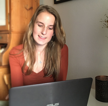 Eine lächelnde Frau arbeitet an einem Tisch an einem Laptop. Sie trägt ein rostfarbenes Hemd. Innenaufnahme.